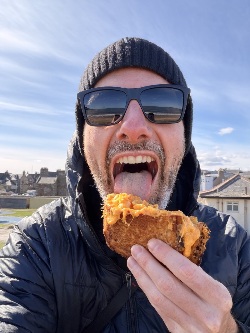 James eating a Mac 'n' Cheese sandwich on the beach in St Andrews, Scotland